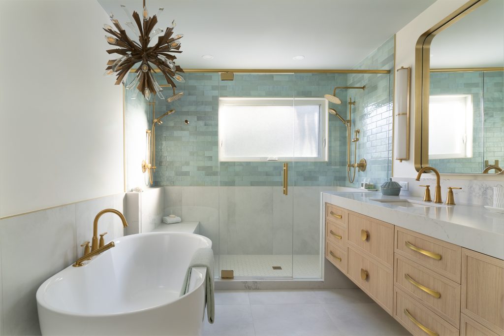 An amazing bathroom with large walk-in shower with light blue and white tiles, soaker tub and chandelier, and light wood-grained vanity with gold hardware.