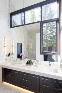 A dark wood vanity with chrome hardware, large white countertop, large mirror, windows, and long wall sconce light.