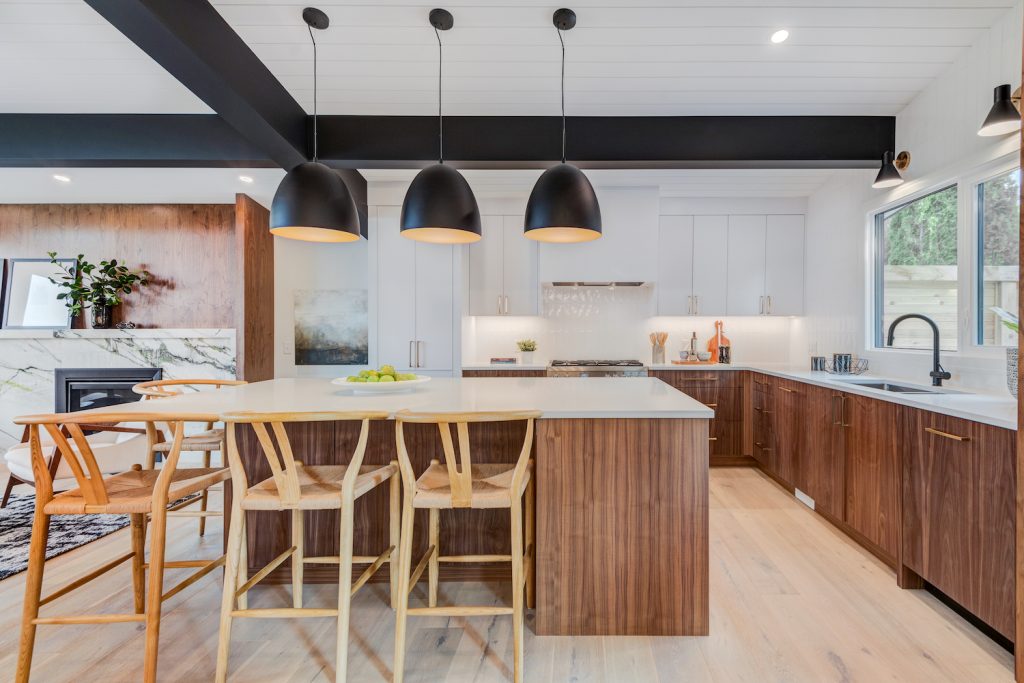 Large kitchen with wood-grain cabinets, white stone countertops, kitchen island with seating, large black pendant lights, and open black wood ceiling beams.