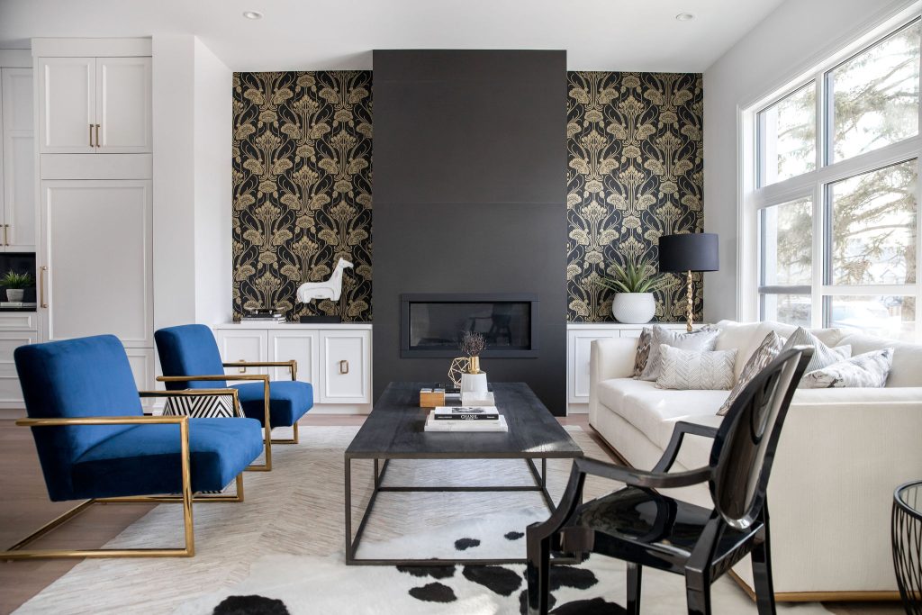 A modern designer living room with a black fireplace, lower white cabinets, elegant gold and black wallpaper, royal blue chairs and a white sofa.
