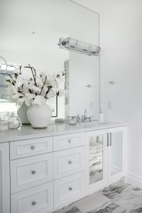 A beautiful bathroom vanity with large mirror, lighting, white cabinetry with built in mirrors, and a white vase with white flowers.