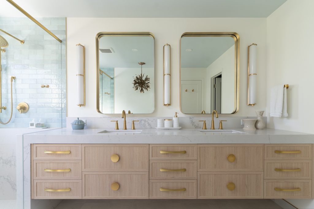 A modern bathroom vanity with light wood grain cabinetry and copper handles, double sinks, and two mirrors.