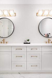 A beautiful bathroom vanity with two sinks, white cabinets with gold hardware, two black round mirrors, gold faucets and gold lighting.