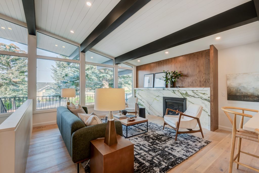 A newly renovated retro living room with a marble fireplace with wood panelling, light wood flooring, and a vaulted ceiling with large dark brown beams.