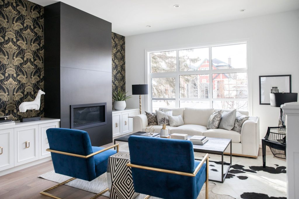 A modern designer living room with a black fireplace, white cabinets, elegant gold and black wallpaper, a large white sofa and royal blue chairs.