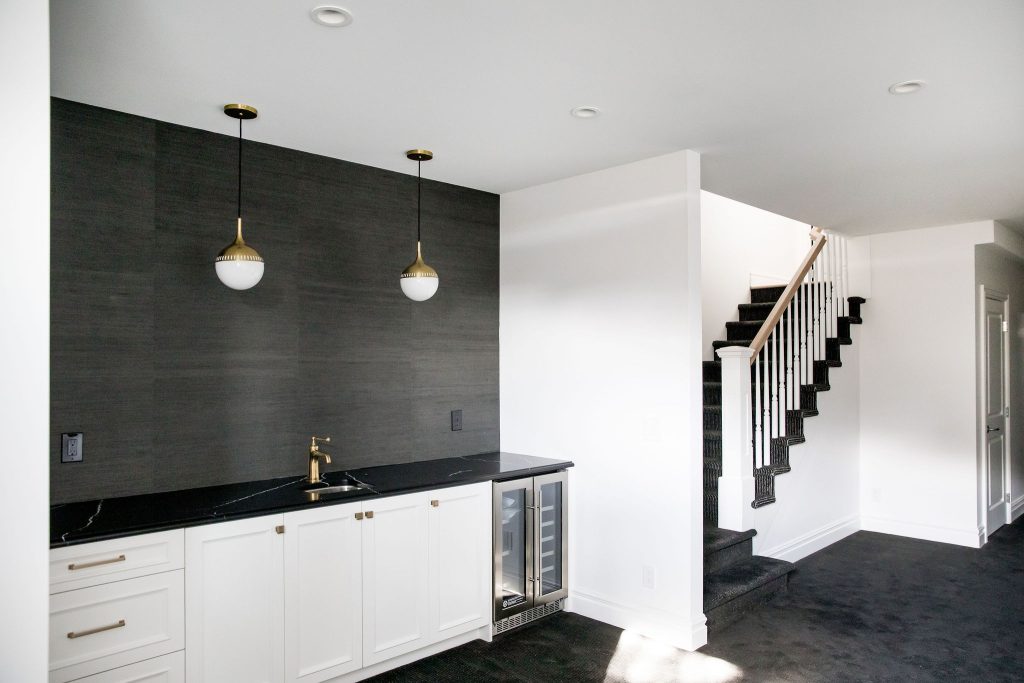 A modern basement kitchen with a sink, bar fridge, white cabinets, and a black feature wall.