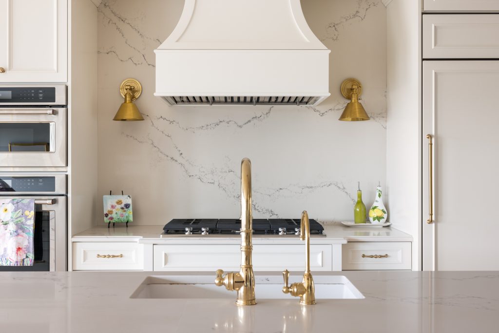A stunning gold kitchen sink faucet in an island across from a gas stove with a hood fan, gold accent lights and marble backsplash.