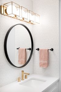 A bathroom vanity with a round black mirror, white countertop, gold faucet, gold vanity lighting, and black towel holder.