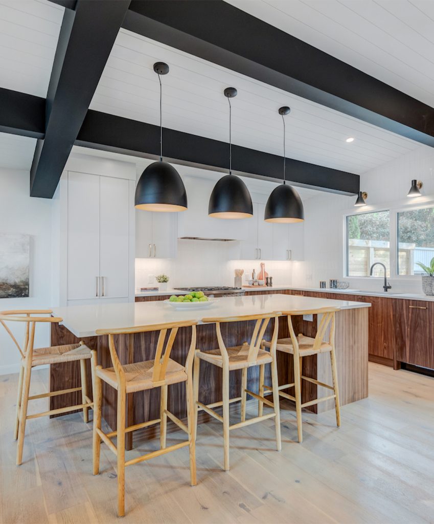 A modern white kitchen with wood cabinetry, large black ceiling beams and black pendant lights above the island.