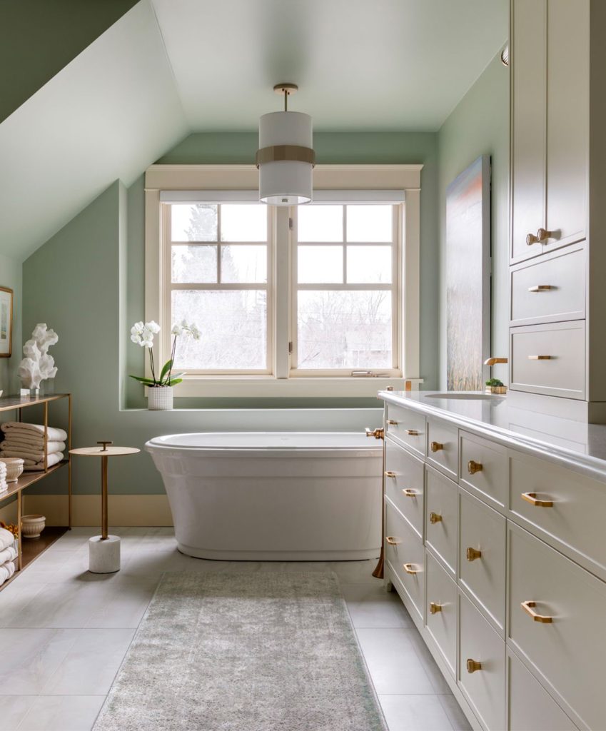 A newly renovated bathroom with, softly lit sea green walls, cream cabinetry and a stand alone tub.